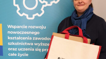 Uśmiechnięta osoba trzyma beżową torbę z kolorowymi uchwytami, stojąc obok niebieskiego znaku z ikonami kół zębatych i polskim tekstem o wsparciu kształcenia zawodowego i ustawicznego. Na znaku i torbie widnieją logotypy Polski, UE i województwa opolskiego.