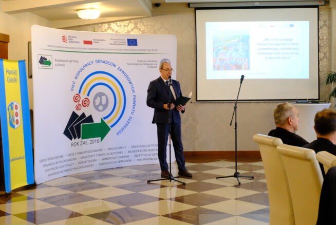 Mężczyzna w garniturze stoi przy mikrofonie z otwartą teczką, przemawiając na imprezie wewnętrznej. Za nim znajduje się baner z logo, tekstem i symbolami oraz ekran projektora wyświetlający slajd prezentacji z tekstem. Podłoga ma wzór w brązowo-białą kratkę.