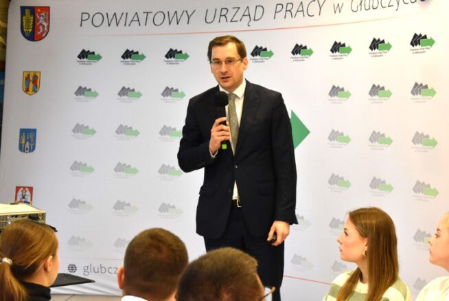 Mężczyzna w granatowym garniturze i okularach trzyma mikrofon, przemawiając do siedzącej publiczności. Za nim transparent z polskim tekstem: POWIATOWY URZĄD PRACY W GŁUBCZYCACH i kilkoma logotypami. Czworo młodych ludzi stoi przed nim, słuchając uważnie.