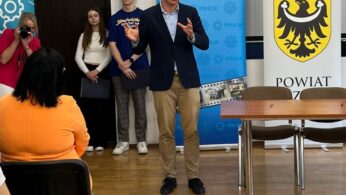 Mężczyzna w ciemnej marynarce przemawia przed grupą, gestykulując rękami. Za nim stoi troje młodych ludzi i fotograf. Na ścianie wiszą transparenty policji i powiatu. Kilka osób siedzi naprzeciwko mówcy w jasno oświetlonym pomieszczeniu.