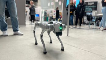 Mały, szary pies-robot ze świecącymi zielonymi oczami stoi na betonowej podłodze w pomieszczeniu. W tle widać kilka osób w zwykłych ubraniach, które obserwują wyświetlacze i rozmawiają w wypełnionej stoiskami przestrzeni wystawowej.
