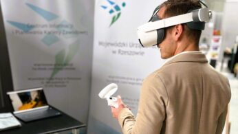 Mężczyzna w beżowej marynarce korzysta z białego zestawu słuchawkowego VR i kontrolera. Stoi w pobliżu laptopa i dwóch wysokich banerów z polskim tekstem i logo w nowoczesnym wnętrzu, prawdopodobnie na wydarzeniu technologicznym lub wystawie.
