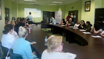 Grupa dorosłych siedzi wokół dużego stołu w kształcie litery U w sali konferencyjnej, słuchając prezentacji. Jedna osoba stoi przy ekranie projektora, podczas gdy inni stoją przodem, niektórzy z papierami lub notatnikami. Pokój jest jasny, a na ścianach wiszą plakaty i banery.