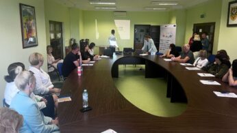 Grupa ludzi, zarówno mężczyzn, jak i kobiet, siedzi wokół dużego stołu konferencyjnego w kształcie litery U w jasnej sali konferencyjnej, słuchając dwóch prezenterów z przodu w pobliżu ekranu projektora. Na stołach leżą papiery, butelki z wodą i notatniki.