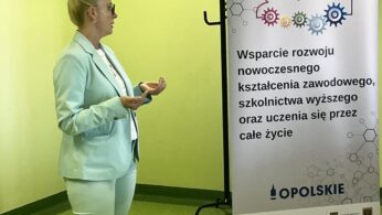 Kobieta w jasnozielonym garniturze stoi i gestykuluje, przemawiając obok baneru z polskim tekstem o nowoczesnej edukacji i uczeniu się przez całe życie. Ściana w tle jest jasnozielona, a podłoga zielona. Baner zawiera logo i kolorowe ludzkie ikony.