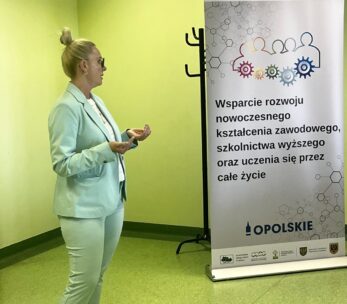 Kobieta w jasnozielonym garniturze stoi i gestykuluje, przemawiając obok baneru z polskim tekstem o nowoczesnej edukacji i uczeniu się przez całe życie. Ściana w tle jest jasnozielona, a podłoga zielona. Baner zawiera logo i kolorowe ludzkie ikony.
