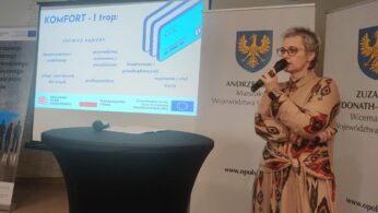 Kobieta trzymająca mikrofon stoi obok ekranu projektora prezentującego slajd zatytułowany KOMFORT - I trop. Slajd zawiera listę tematów związanych z karierą w języku polskim. Za nią znajdują się banery z oficjalnymi polskimi godłami i logo organizacji wspierających.