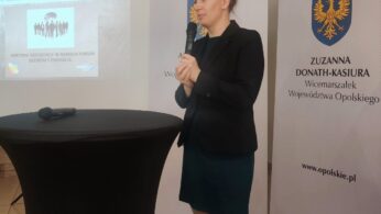 Kobieta w ciemnej marynarce i turkusowej spódnicy stoi, trzymając mikrofon, przemawiając przy wysokim okrągłym stole. Za nią znajdują się banery z herbem i nazwą Zuzanna Donath-Kasiura w języku polskim oraz projektor wyświetlający slajd ze spotkania biznesowego.