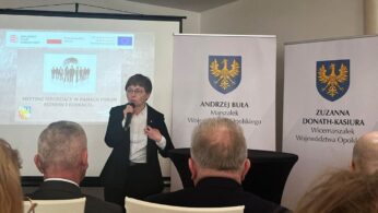 Kobieta w garniturze przemawia do mikrofonu na podium przed publicznością. Za nią znajdują się banery z herbami i nazwiskami polskiego rządu oraz ekran projektora wyświetlający slajd z forum. Na pierwszym planie widoczne są plecy publiczności.
