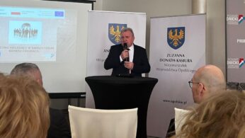 Mężczyzna w garniturze przemawia na podium z mikrofonem. Za nim znajdują się banery z herbem rządu i polskim tekstem. Na pierwszym planie siedzą ludzie. Na ekranie wyświetlany jest slajd z logo i polskim tekstem dotyczącym forum biznesu i edukacji.