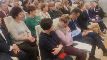 Duża grupa dorosłych w formalnych strojach siedzi blisko siebie na białych krzesłach w dobrze oświetlonym pomieszczeniu, uważnie obserwując lub słuchając niewidocznego mówcy. Niektóre osoby trzymają notatniki lub tablety; atmosfera wydaje się poważna i profesjonalna, prawdopodobnie jest to konferencja lub spotkanie.