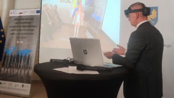 Mężczyzna w garniturze w goglach VR stoi przy okrągłym stole z laptopem, gestykulując w kierunku projekcji przedstawiającej układ mięśniowy człowieka. W tle widoczne są banery z polskim tekstem i flagami UE, co sugeruje wydarzenie zawodowe lub edukacyjne.