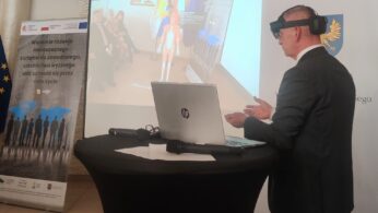 Mężczyzna w garniturze nosi okulary VR i gestykuluje rękami, stojąc przy okrągłym stole z laptopem. Za nim na ekranie widać wirtualny model anatomiczny i siedzących ludzi. W pobliżu wyświetlane są banery z polskim tekstem i logo.