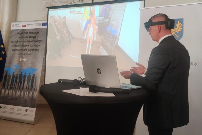 Mężczyzna w garniturze nosi okulary VR i gestykuluje rękami, stojąc przy okrągłym stole z laptopem. Za nim na ekranie widać wirtualny model anatomiczny i siedzących ludzi. W pobliżu wyświetlane są banery z polskim tekstem i logo.