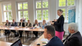 Kobieta stoi i przemawia do grupy siedzącej wokół stołu konferencyjnego w jasnym pomieszczeniu z dużymi oknami. Uczestnicy uważnie słuchają; na stole leżą laptopy, papiery i szklanki z wodą. W tle widoczny jest baner z kołami zębatymi.