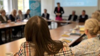 Grupa ludzi siedzi wokół stołu konferencyjnego w jasnym pomieszczeniu, skupiając się na kobiecie stojącej i przemawiającej z przodu. Na stole leżą papiery, książki i filiżanki do kawy. W tle widoczny jest ekran projekcyjny i baner.
