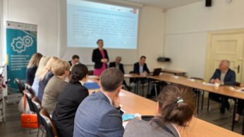 Grupa ludzi siedzi wokół stołu konferencyjnego w jasnej sali konferencyjnej, podczas gdy kobieta stoi i prezentuje w pobliżu ekranu projekcyjnego. Uczestnicy uważnie słuchają, a na stole widoczne są dokumenty i laptopy. W rogu stoi duży baner.