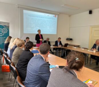 Grupa ludzi siedzi wokół stołu konferencyjnego w jasnej sali konferencyjnej, podczas gdy kobieta stoi i prezentuje w pobliżu ekranu projekcyjnego. Uczestnicy uważnie słuchają, a na stole widoczne są dokumenty i laptopy. W rogu stoi duży baner.
