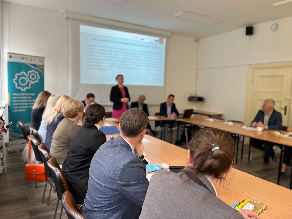 Grupa ludzi siedzi wokół stołu konferencyjnego w jasnej sali konferencyjnej, podczas gdy kobieta stoi i prezentuje w pobliżu ekranu projekcyjnego. Uczestnicy uważnie słuchają, a na stole widoczne są dokumenty i laptopy. W rogu stoi duży baner.