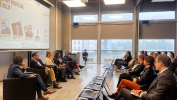 Panel sześciu osób siedzi na scenie przed publicznością w nowoczesnym pomieszczeniu z dużymi oknami. Na ekranie projektora wyświetlane są zdjęcia i nazwiska. Uczestnicy siedzą w rzędach, twarzą do mówców. Naturalne światło wypełnia pomieszczenie, a jedna osoba stoi z tyłu.