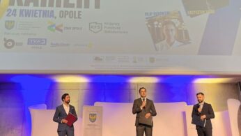 Trzech mężczyzn w garniturach stoi na scenie z mikrofonami podczas Festiwalu Kariery w Opolu. Za nimi znajduje się duży ekran wyświetlający szczegóły wydarzenia, zdjęcia gości i logo. Publiczność jest częściowo widoczna na pierwszym planie.