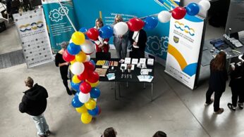 Stoisko na targach kariery z czterema osobami za stołem wyświetlającym broszury i ulotki. Kolorowe czerwone, białe, niebieskie i żółte balony tworzą łuk. Uczestnicy w swobodnych strojach przechodzą obok lub wchodzą w interakcje. Duże banery z logo i tekstem stoją za stoiskiem.