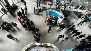 Tętniące życiem wydarzenie pod dachem z ludźmi spacerującymi i gromadzącymi się w kolejkach przy stoiskach i stołach. Niektóre stoiska mają kolorowe balony i wyświetlacze informacyjne. Zdjęcie zostało zrobione z góry, pokazując przestronną halę z dużymi oknami i różnymi sekcjami wystawowymi.