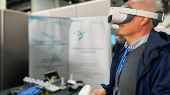Starszy mężczyzna ubrany w kurtkę i niebieską smycz używa białego zestawu słuchawkowego wirtualnej rzeczywistości i trzyma kontrolery VR. Stoi przed stoiskiem wystawowym z niewyraźnymi banerami i sprzętem technicznym na stole w tle.