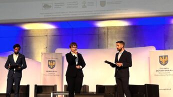 Trzy osoby w formalnych strojach stoją na scenie z mikrofonami i dokumentami. Pośrodku przemawia kobieta otoczona przez dwóch mężczyzn. Za nimi znajdują się puste krzesła i banery z logo herbu. Tło jest oświetlone niebieskim światłem.