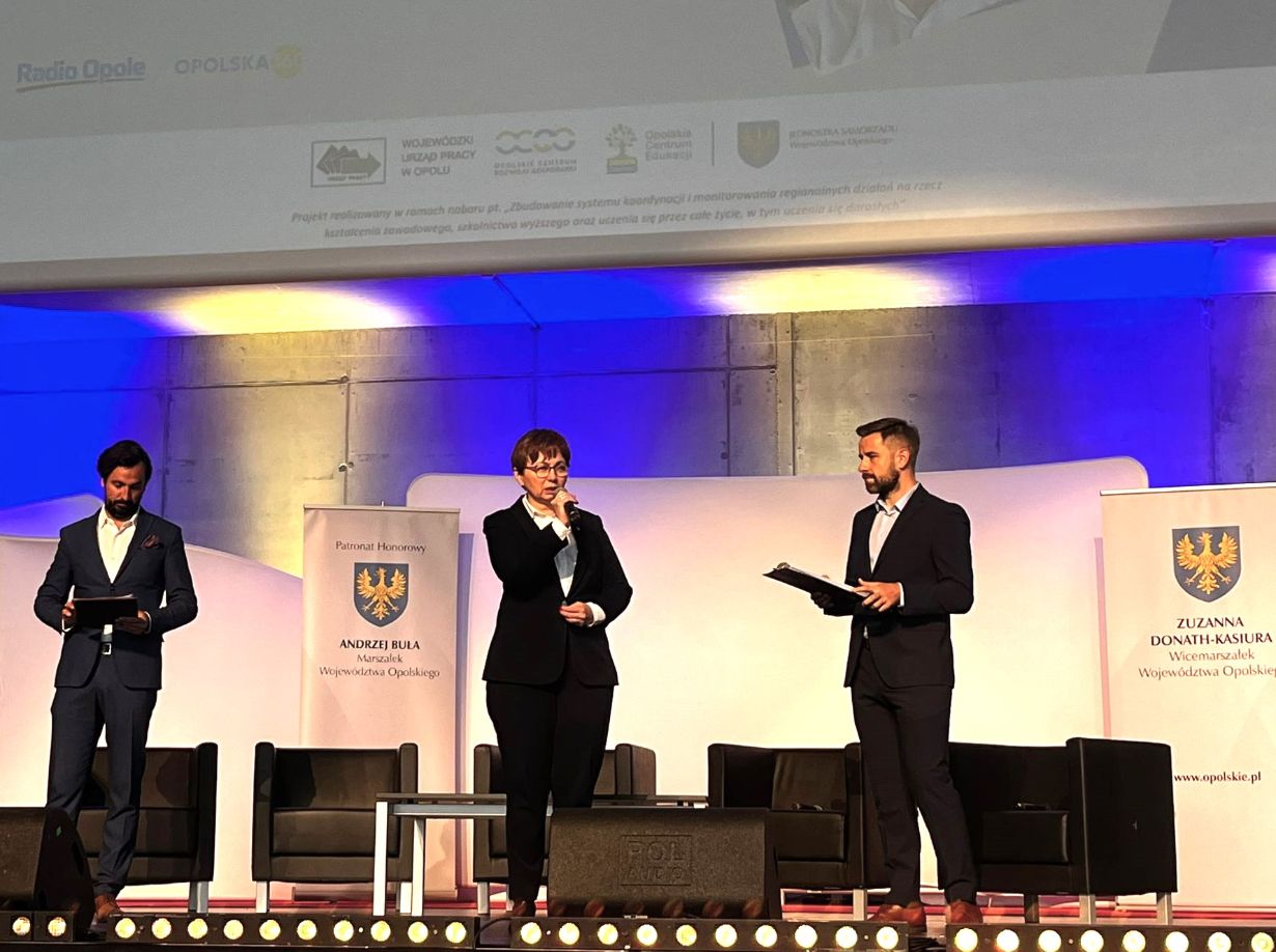 Trzy osoby w formalnych strojach stoją na scenie z mikrofonami i dokumentami. Pośrodku przemawia kobieta otoczona przez dwóch mężczyzn. Za nimi znajdują się puste krzesła i banery z logo herbu. Tło jest oświetlone niebieskim światłem.