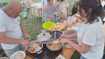 Mężczyzna i kobieta gotują jedzenie przy stole na świeżym powietrzu z patelniami na kuchence, przygotowując mięso i placki. W pobliżu stoi kilka kobiet i dziewcząt, niektóre we wzorzystych sukienkach. Stół jest pełen jedzenia, misek i przyborów, pod namiotem na trawiastym terenie.