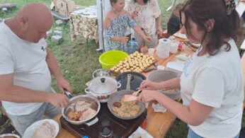 Dwoje dorosłych gotuje jedzenie na przenośnych kuchenkach przy stole pod namiotem, smażąc mięso i placki. Kilka kobiet i dziewcząt stoi w pobliżu, niektóre uśmiechnięte, z wypiekami i naczyniami na stole. W tle zielona trawa i przedmioty piknikowe.