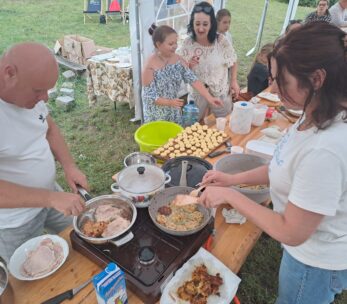 Dwoje dorosłych gotuje jedzenie na przenośnych kuchenkach przy stole pod namiotem, smażąc mięso i placki. Kilka kobiet i dziewcząt stoi w pobliżu, niektóre uśmiechnięte, z wypiekami i naczyniami na stole. W tle zielona trawa i przedmioty piknikowe.