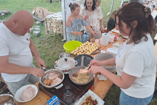 Dwoje dorosłych gotuje jedzenie na przenośnych kuchenkach przy stole pod namiotem, smażąc mięso i placki. Kilka kobiet i dziewcząt stoi w pobliżu, niektóre uśmiechnięte, z wypiekami i naczyniami na stole. W tle zielona trawa i przedmioty piknikowe.