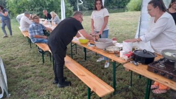 Ludzie przygotowują jedzenie na drewnianych ławkach pod białym namiotem z baldachimem. Niektórzy siedzą, jedzą i rozmawiają, podczas gdy inni stoją przy stole mieszając składniki, otoczeni przyborami kuchennymi. Scena jest swobodna i osadzona na trawie z drzewami w tle.