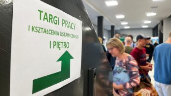 Napis w języku polskim na ścianie z zieloną strzałką kieruje na piętro z napisem Targi Pracy i Kształcenia Ustawicznego I Piętro. W tle kilka osób idzie dobrze oświetlonym korytarzem.