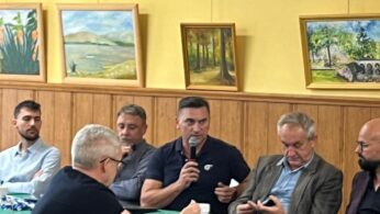 Grupa mężczyzn siedzi wokół zielonego stołu i dyskutuje; jeden z nich mówi do mikrofonu. Na żółtej ścianie za nimi wiszą obrazy przedstawiające krajobrazy i kwiaty. Na stole leży kilka kubków, papiery i telefon.