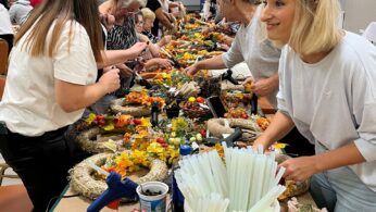 Pokój pełen ludzi, głównie kobiet, gromadzi się wokół długich stołów pokrytych kolorowymi kwiatami, słomianymi wieńcami i artykułami rzemieślniczymi. Są uśmiechnięci i skupieni, tworząc kompozycje kwiatowe. Atmosfera jest żywa i twórcza.