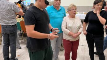 Młody mężczyzna noszący zestaw VR stoi w pokoju, trzymając kontrolery. Kilku starszych dorosłych i młoda kobieta obserwują go z zainteresowaniem. Grupa znajduje się w pomieszczeniu, stojąc na jasnej drewnianej podłodze; w tle znajdują się stoły z jedzeniem i dekoracjami.