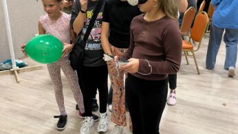 Grupa czterech dziewcząt stoi w pomieszczeniu; jedna nosi biały zestaw VR, podczas gdy pozostałe ją obserwują. Są ubrane swobodnie, a jedna z nich trzyma zielony balon. W tle jasno oświetlonego pokoju widać więcej osób i pomarańczowe krzesła.
