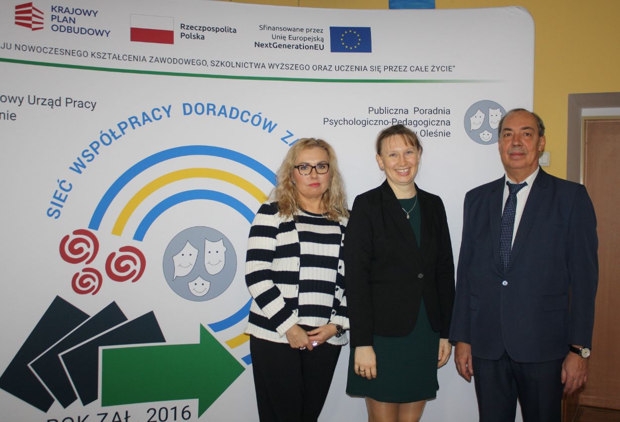 Trzy dorosłe osoby, dwie kobiety i jeden mężczyzna, stoją uśmiechnięte w strojach biznesowych przed wyświetlaczem z polskim tekstem, symbolami i logo programów edukacyjnych i unijnych. Tło zawiera niebieskie strzałki, obrazy sieci i oficjalne logo.