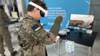 Osoba w mundurze maskującym korzysta z zestawu słuchawkowego i kontrolera wirtualnej rzeczywistości. Za nimi znajduje się stół z monitorem, zestawem VR i materiałami informacyjnymi. Inna osoba w mundurze jest częściowo widoczna w tle obok niebieskiej tablicy.