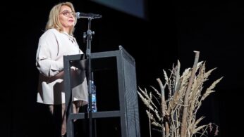 Kobieta o blond włosach i w okularach przemawia z podium na ciemnej scenie. Ma na sobie jasną bluzkę i spodnie. Do podium przymocowany jest mikrofon, a na dolnej półce stoi butelka z wodą. Suszone rośliny są ułożone dekoracyjnie na pierwszym planie.