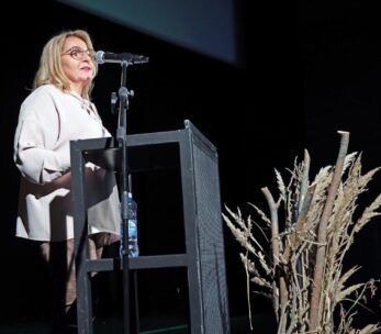 Kobieta o blond włosach i w okularach przemawia z podium na ciemnej scenie. Ma na sobie jasną bluzkę i spodnie. Do podium przymocowany jest mikrofon, a na dolnej półce stoi butelka z wodą. Suszone rośliny są ułożone dekoracyjnie na pierwszym planie.