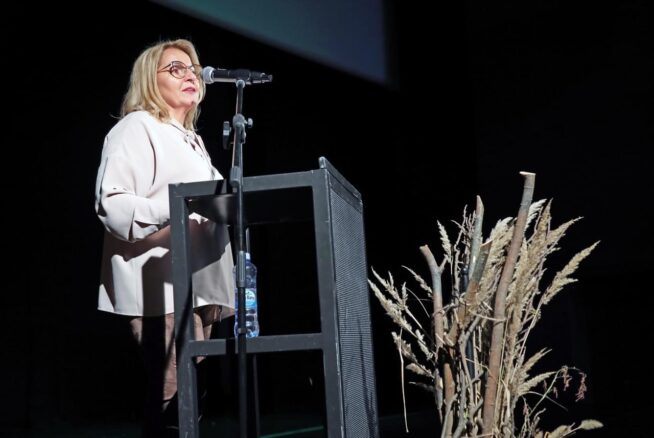 Kobieta o blond włosach i w okularach przemawia z podium na ciemnej scenie. Ma na sobie jasną bluzkę i spodnie. Do podium przymocowany jest mikrofon, a na dolnej półce stoi butelka z wodą. Suszone rośliny są ułożone dekoracyjnie na pierwszym planie.