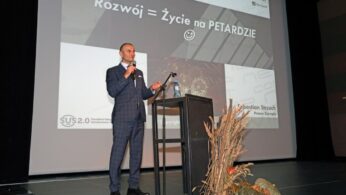 Mężczyzna w kraciastym garniturze stoi na scenie i mówi do mikrofonu, trzymając w ręku pilota. Za nim na dużym ekranie wyświetlana jest polska prezentacja Rozwój = Życie na PETARDZIE :), z logotypami i nazwiskiem Sebastiana Sierżęgi. Z przodu znajduje się rustykalna dekoracja.