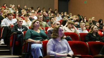 Widzowie w różnym wieku siedzą uważnie na czerwonych fotelach w dużym audytorium. Większość osób zwrócona jest do przodu, skupiając się na niewidocznej scenie lub ekranie. Pomieszczenie jest dobrze oświetlone, a tłum wydaje się zaangażowany i zamyślony.