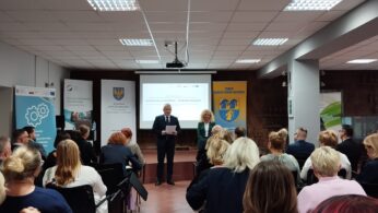 Mężczyzna w garniturze i kobieta stoją z przodu sali, przemawiając do publiczności siedzącej w rzędach. Za nimi znajduje się ekran projektora wyświetlający prezentację, z banerami i plakatami po obu stronach pokoju pod jasnymi światłami sufitowymi.