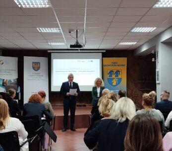 Mężczyzna w garniturze i kobieta stoją z przodu sali, przemawiając do publiczności siedzącej w rzędach. Za nimi znajduje się ekran projektora wyświetlający prezentację, z banerami i plakatami po obu stronach pokoju pod jasnymi światłami sufitowymi.