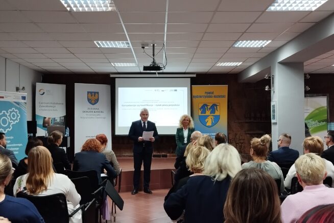Mężczyzna w garniturze i kobieta stoją z przodu sali, przemawiając do publiczności siedzącej w rzędach. Za nimi znajduje się ekran projektora wyświetlający prezentację, z banerami i plakatami po obu stronach pokoju pod jasnymi światłami sufitowymi.